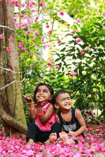laughing boy and girl sitting on pink flower petals near tree